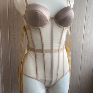 May include: A beige corset-style bodysuit with a structured bust and sheer panels. The garment features adjustable straps and ribbon ties, displayed on a mannequin. The color palette is neutral, with a satin-like finish on the bust.
