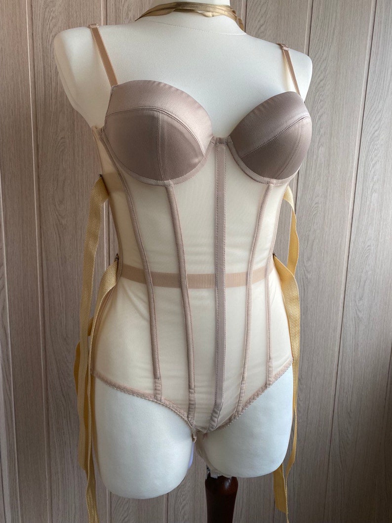 May include: A beige corset-style bodysuit with a structured bust and sheer panels. The garment features adjustable straps and ribbon ties, displayed on a mannequin. The color palette is neutral, with a satin-like finish on the bust.