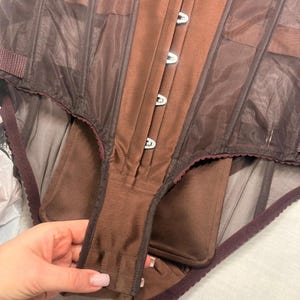 May include: A brown vintage corset with a structured design. The corset features a central panel with metal clasps, sheer mesh side panels, and a dark burgundy trim. The garment is held by a hand.
