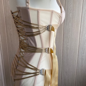 May include: A beige and cream-colored corset with a structured design. The corset features a lace-up back, multiple straps, and metal clasps. The garment is displayed on a mannequin, showcasing its shape and details. The overall aesthetic is elegant and refined.