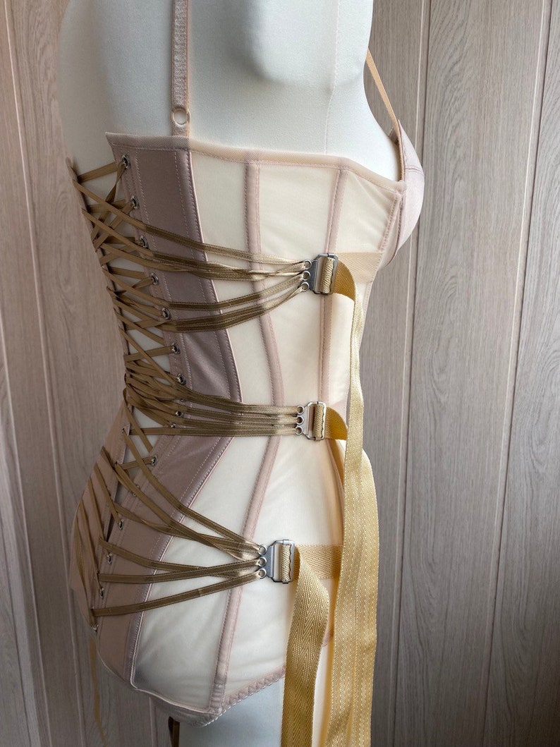 May include: A beige and cream-colored corset with a structured design. The corset features a lace-up back, multiple straps, and metal clasps. The garment is displayed on a mannequin, showcasing its shape and details. The overall aesthetic is elegant and refined.