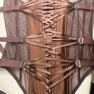 May include: A brown corset with a satin finish and mesh panels. The corset features a lace-up back and adjustable straps with silver-colored metal buckles. The garment has a structured silhouette and a built-in bottom.