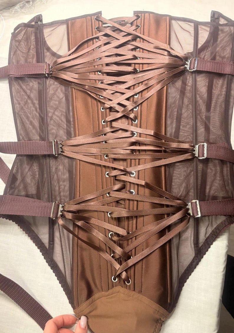 May include: A brown corset with a satin finish and mesh panels. The corset features a lace-up back and adjustable straps with silver-colored metal buckles. The garment has a structured silhouette and a built-in bottom.