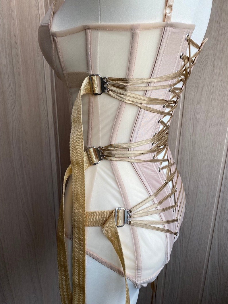 May include: A beige corset with a structured design, featuring vertical boning and a lace-up back. The corset has gold-colored straps and buckles, and is displayed on a mannequin. The garment is made of a combination of sheer and opaque fabrics.