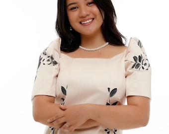 May include: A light beige dress with a square neckline and short sleeves. The dress features a floral design in black, with the flowers and leaves painted on the front and sleeves. A pearl necklace is worn.