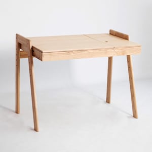 Puede incluir: Un escritorio de madera clara con una superficie rectangular de diseño dividido, que incluye una pequeña abertura circular en el lado derecho. El escritorio se apoya sobre cuatro patas inclinadas con orificios visibles para ajustar la altura.