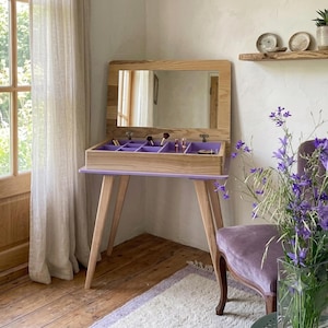 Op de afbeelding: Een kaptafel van licht hout met een opklapbare spiegel en een paars opbergvak aan de binnenkant dat is verdeeld in meerdere secties. De kaptafel heeft vier taps toelopende houten poten en staat in een kamer met een raam en een paarse stoel.