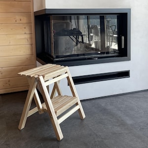 Puede incluir: Un pequeño taburete plegable de madera de dos escalones. El taburete está hecho de madera de color claro y tiene un diseño simple y funcional. Se coloca frente a una chimenea moderna con marco negro y puerta de vidrio.