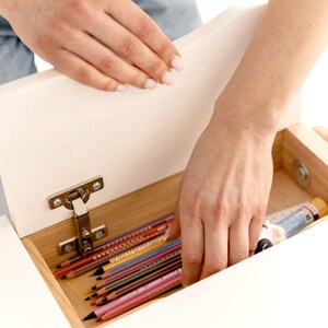 May include: A white wooden desk drawer is opened to reveal a collection of colored pencils and art supplies. The drawer is attached with a silver hinge. The pencils are various colors, including red, blue, and yellow.