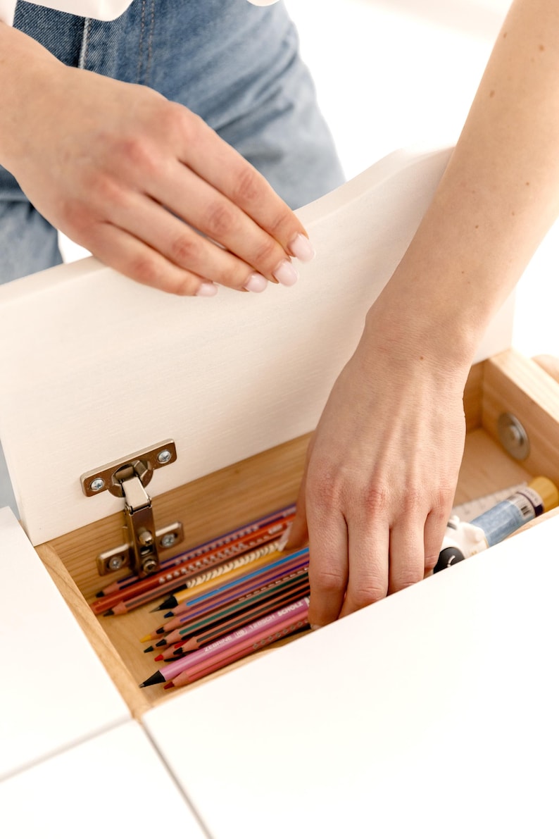 May include: A white wooden desk drawer is opened to reveal a collection of colored pencils and art supplies. The drawer is attached with a silver hinge. The pencils are various colors, including red, blue, and yellow.