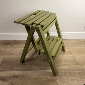 Può includere: Uno sgabello a gradini in legno verde con due gradini. Il gradino superiore ha un piccolo ritaglio al centro.
