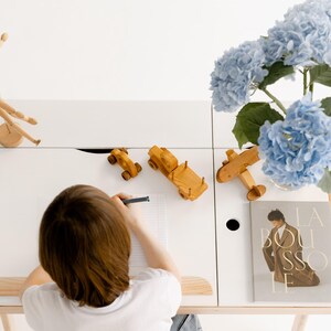 May include: A white desk with wooden accents, featuring wooden toy cars, a plane, and a poseable wooden figure. A vase of blue hydrangeas and a book are also on the desk. A person is drawing on a piece of paper.