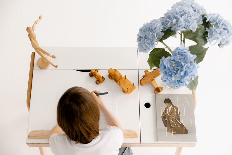 May include: A white desk with wooden accents, featuring wooden toy cars, a plane, and a poseable wooden figure. A vase of blue hydrangeas and a book are also on the desk. A person is drawing on a piece of paper.