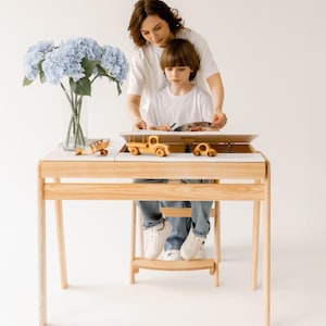 Pode incluir: Um conjunto de mesa e cadeira de madeira para crianças com tampo branco e estrutura de madeira clara. Uma criança está sentada à mesa com um adulto, desenhando em um papel. Há carrinhos de brinquedo e um avião na mesa.