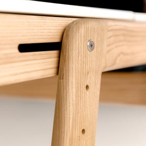 May include: Close-up of a wooden table leg and frame joint. The light-colored wood has a natural grain and is joined with a metal fastener. The design features a black slot and several drilled holes.