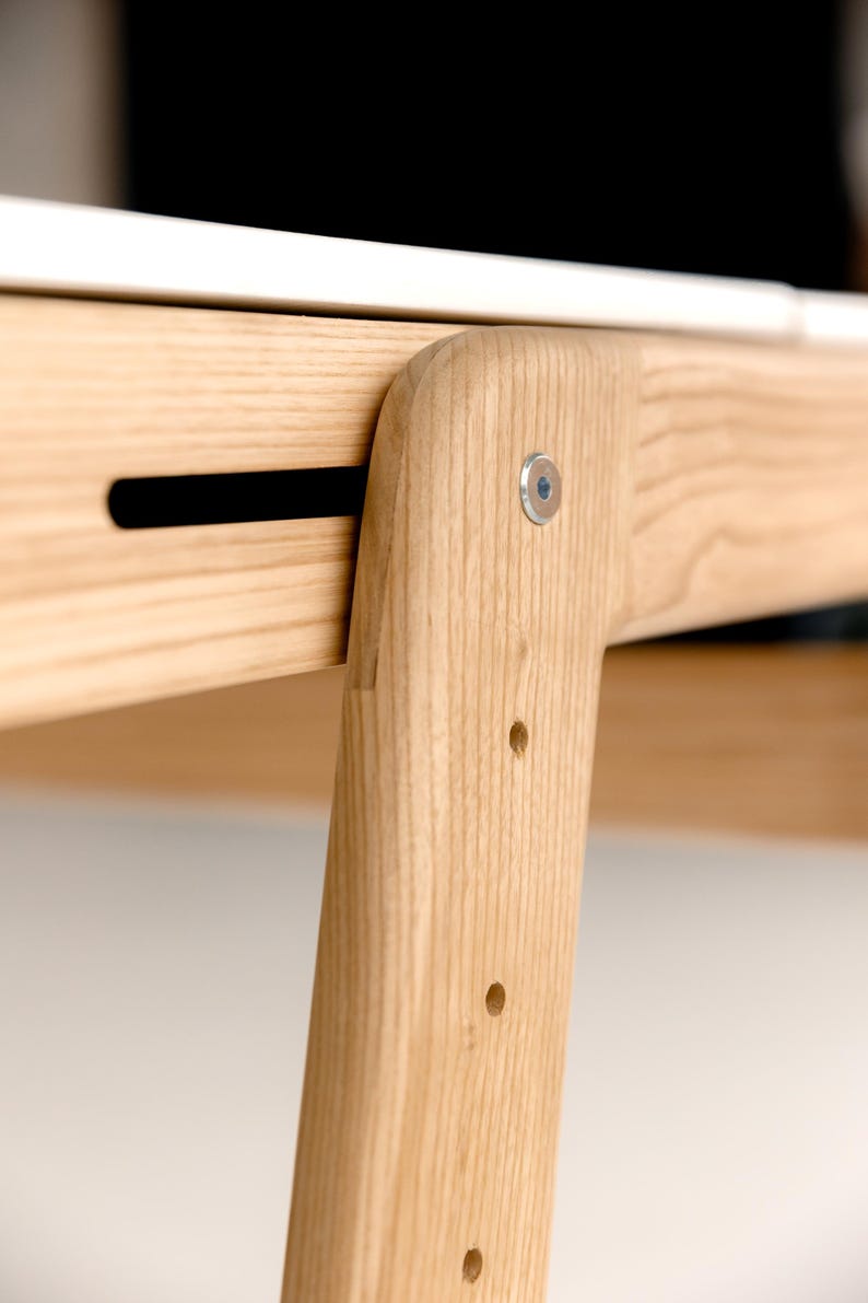 May include: Close-up of a wooden table leg and frame joint. The light-colored wood has a natural grain and is joined with a metal fastener. The design features a black slot and several drilled holes.