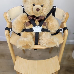 May include: A light brown teddy bear wearing a black and brown safety harness in a wooden high chair with a step stool.