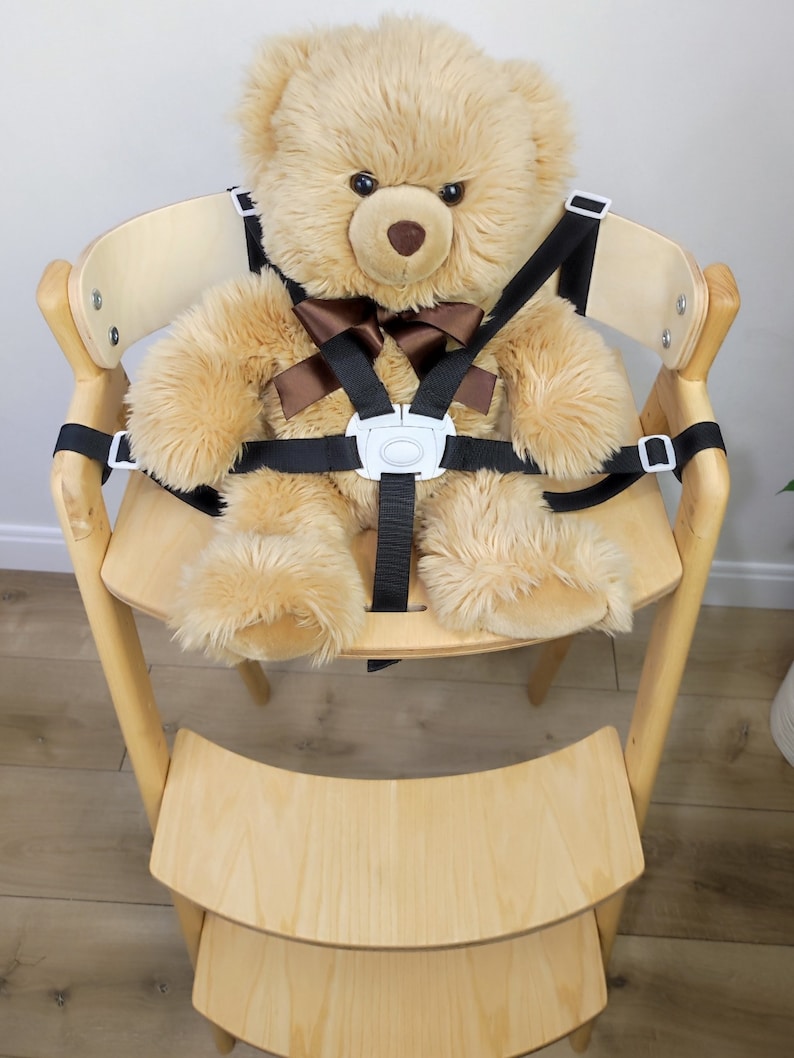 May include: A light brown teddy bear wearing a black and brown safety harness in a wooden high chair with a step stool.