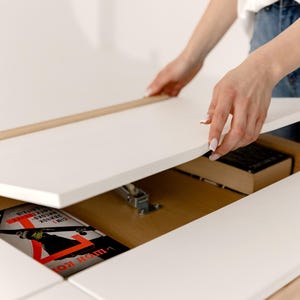 May include: A white table with a hidden compartment is being opened, revealing a book and other items. The table has a light wood trim and a smooth, clean surface. The book has a red and black cover.