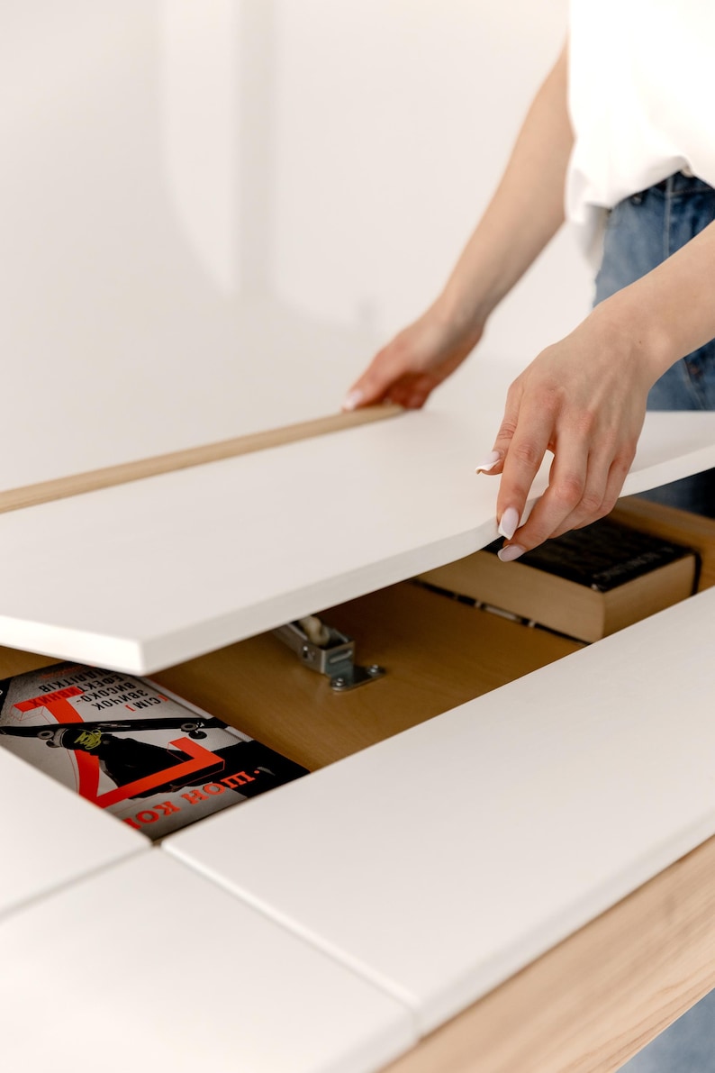 May include: A white table with a hidden compartment is being opened, revealing a book and other items. The table has a light wood trim and a smooth, clean surface. The book has a red and black cover.
