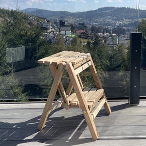Può includere: Un piccolo sgabello in legno pieghevole a due gradini. Lo sgabello è realizzato in legno chiaro e presenta un design a doghe sui gradini e sulla parte superiore. Le gambe sono angolate per la stabilità. Lo sfondo mostra una vista panoramica di montagne e una città.