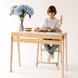 Pode incluir: Uma secretária de madeira clara com um tampo branco, com uma criança a ler. A secretária tem um apoio para os pés embutido. Um avião de brinquedo e um comboio de madeira estão na secretária. Um vaso de flores azuis está à esquerda.