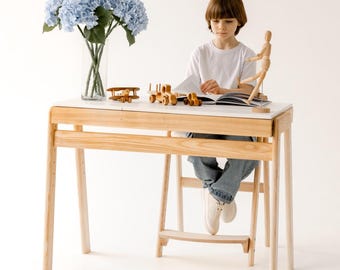 May include: A light wood desk with a white top, featuring a child seated and reading. The desk has a built-in footrest. Wooden toy airplane and train set are on the desk. A vase of blue flowers is on the left.
