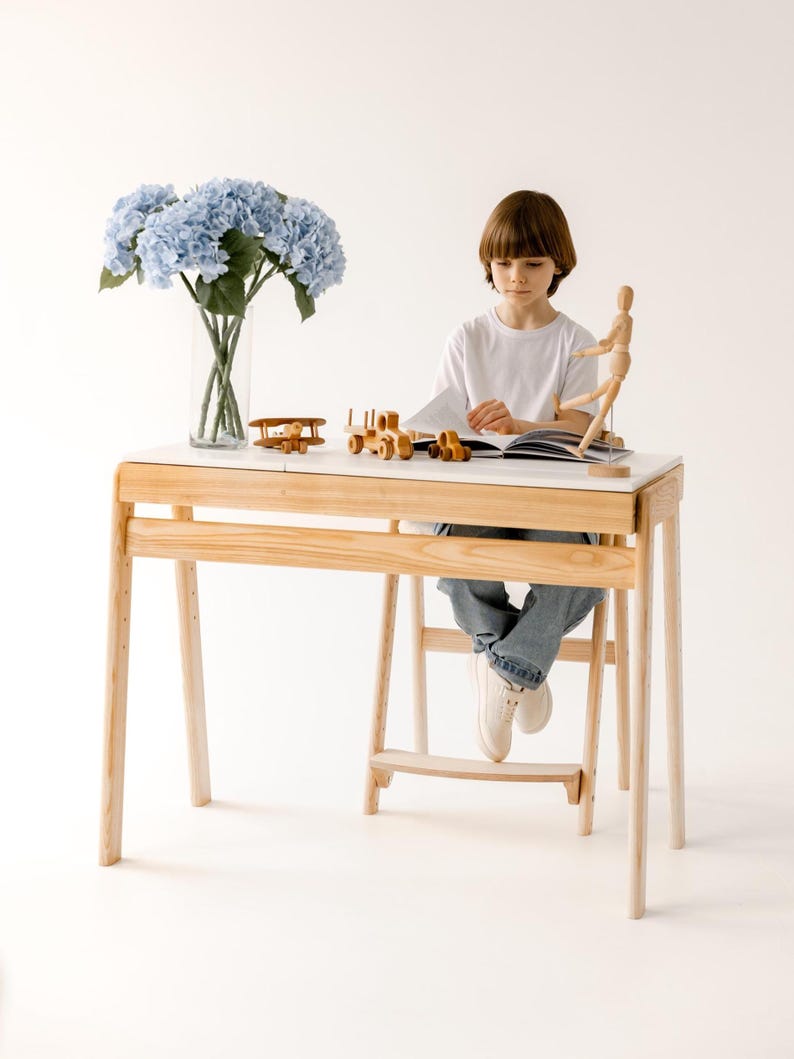 May include: A light wood desk with a white top, featuring a child seated and reading. The desk has a built-in footrest. Wooden toy airplane and train set are on the desk. A vase of blue flowers is on the left.