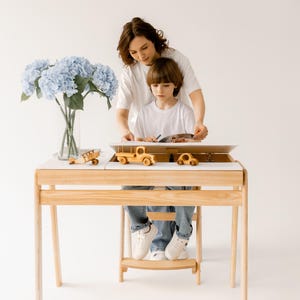 May include: A light wood children's desk with a white top, featuring a built-in storage compartment. The desk holds wooden toy cars and a plane. A vase of blue flowers sits to the side. A person and a child are at the desk.