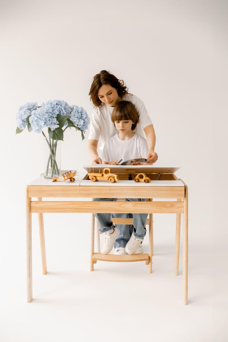 May include: A light wood children's desk with a white top, featuring a built-in storage compartment. The desk holds wooden toy cars and a plane. A vase of blue flowers sits to the side. A person and a child are at the desk.