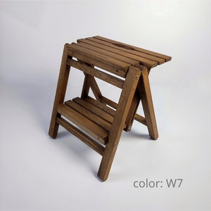Può includere: Uno sgabello a gradini in legno con finitura color legno marrone. Lo sgabello è piegato e ha un piccolo ripiano nella parte inferiore. Il testo "color: W7" è visibile nell'immagine.