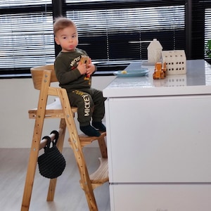 Könnte beinhalten: Ein Holz-Hochstuhl mit einer schwarzen gewebten Tasche, die an der Seite hängt. Ein kleines Kind sitzt auf dem Stuhl und trägt ein grünes Shirt mit dem Aufdruck "Mickey". Der Stuhl steht neben einem weißen Schrank mit zwei Schubladen.