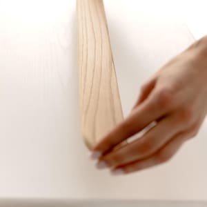 May include: A light-colored wooden furniture component is being handled. The wood grain is visible. The component appears to be part of a larger piece of furniture, possibly a table or desk. The background is a white surface.