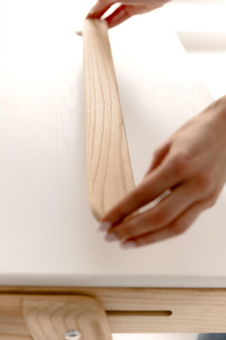 May include: A light-colored wooden furniture component is being handled. The wood grain is visible. The component appears to be part of a larger piece of furniture, possibly a table or desk. The background is a white surface.