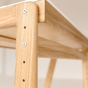May include: Close-up of a wooden table with a white top. The table has a light wood grain finish and is constructed with visible screws and joints. The design is minimalist and modern, suitable for home or office use.
