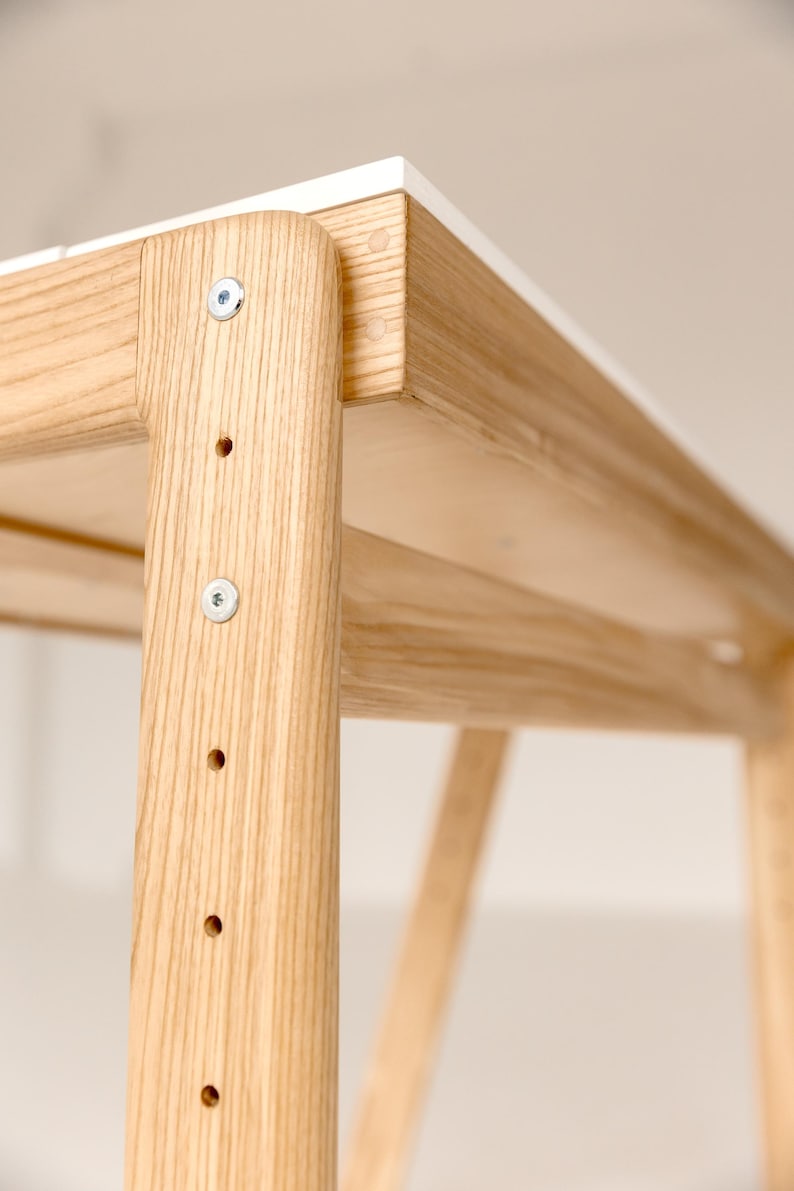 May include: Close-up of a wooden table with a white top. The table has a light wood grain finish and is constructed with visible screws and joints. The design is minimalist and modern, suitable for home or office use.