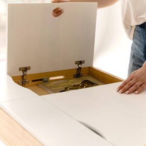 May include: A white wooden desk with a lift-up top, revealing a storage compartment. The desk has a light wood frame and a smooth, white surface. The top is being lifted to show the interior, which contains a book with yellow accents.