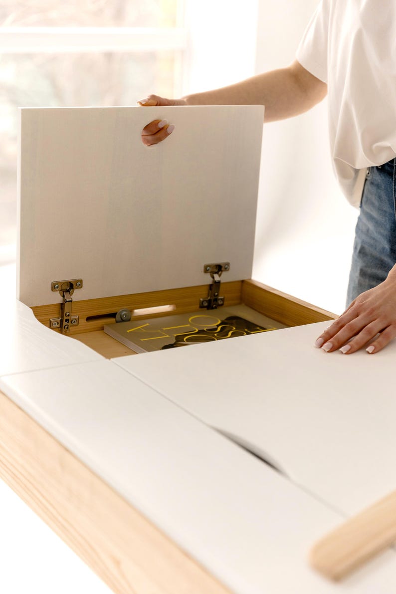 May include: A white wooden desk with a lift-up top, revealing a storage compartment. The desk has a light wood frame and a smooth, white surface. The top is being lifted to show the interior, which contains a book with yellow accents.