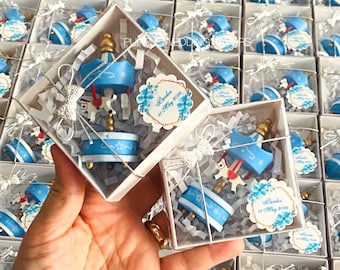 May include: A collection of small, clear boxes filled with blue and white carousel favours. Each box has a white tag with the text "Pandora 11 May 2023".