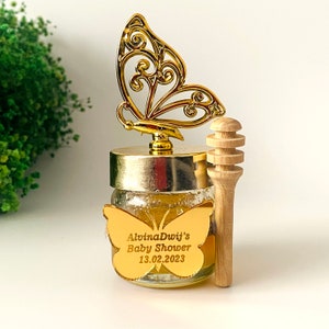 May include: A small glass jar filled with honey, topped with a gold butterfly design and a wooden honey dipper. The jar has a gold label with the text "Alvina Dwij's Baby Shower 13.02.2023".