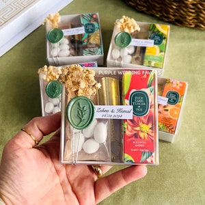 May include: A clear plastic box filled with wedding favors. The box contains a small bottle of cologne, a gold bar, a small bag of white candy, a green wax seal with a leaf design, and a small card with the text "Zehra & Kemal 16.02.2024" and "Eyüp Sabrı Tuncer 1923".