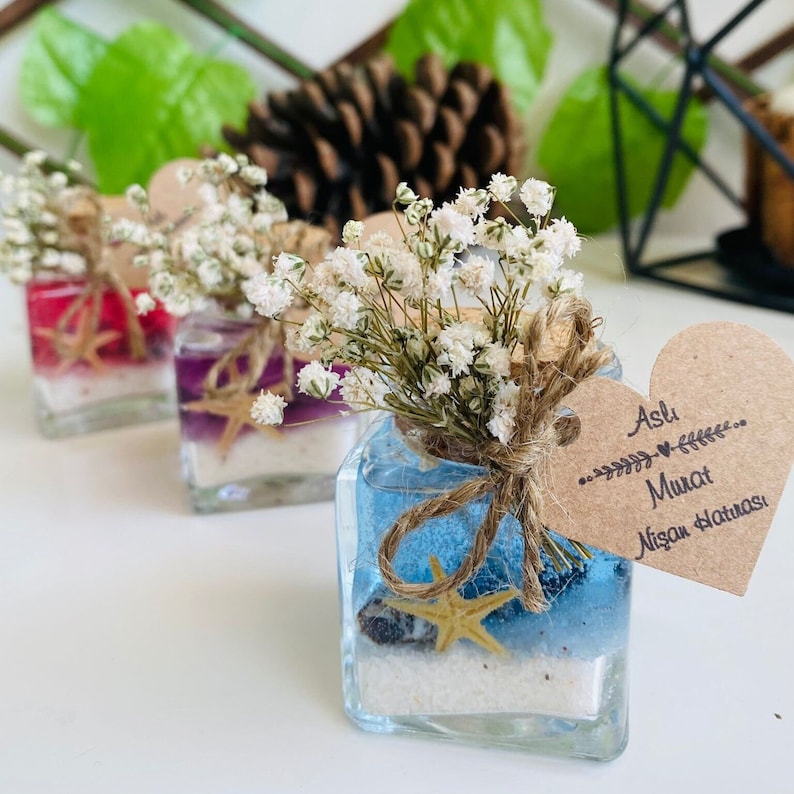 Puede incluir: Peque&ntilde;os frascos de vidrio llenos de gel azul, morado y rojo, arena y conchas marinas. Cada frasco est&aacute; coronado con un peque&ntilde;o ramo de aliento de beb&eacute; blanco y una etiqueta en forma de coraz&oacute;n marr&oacute;n con el texto "Aslı Murat Nişan Hatırası".
