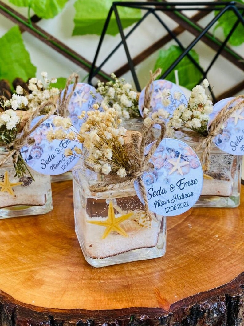 Puede incluir: Peque&ntilde;os frascos de vidrio llenos de gel transparente, arena y conchas marinas. Cada frasco tiene una etiqueta blanca con el texto "Seda & Emre Nisan Hatırası 12.06.2021" y est&aacute; decorado con flores blancas secas y cuerda.