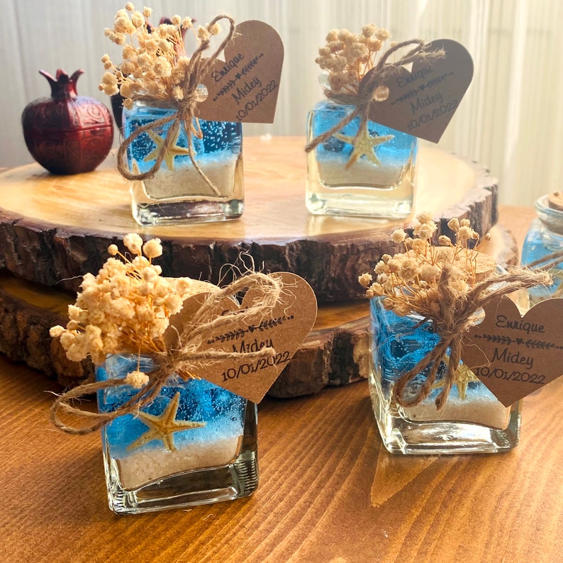 Puede incluir: Cuatro peque&ntilde;os frascos de vidrio con l&iacute;quido azul, arena y una estrella de mar en su interior. Cada frasco est&aacute; decorado con flores secas, cuerda y una etiqueta en forma de coraz&oacute;n con los nombres "Enrique" y "Midey" y la fecha "10/01/2022".