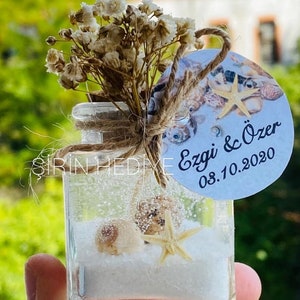 Puede incluir: Un peque&ntilde;o frasco de vidrio lleno de arena blanca y conchas marinas, incluyendo una estrella de mar. El frasco est&aacute; decorado con un lazo de cuerda marr&oacute;n y una etiqueta redonda blanca con el texto "Ezgi & &Ouml;zer 08.10.2020".