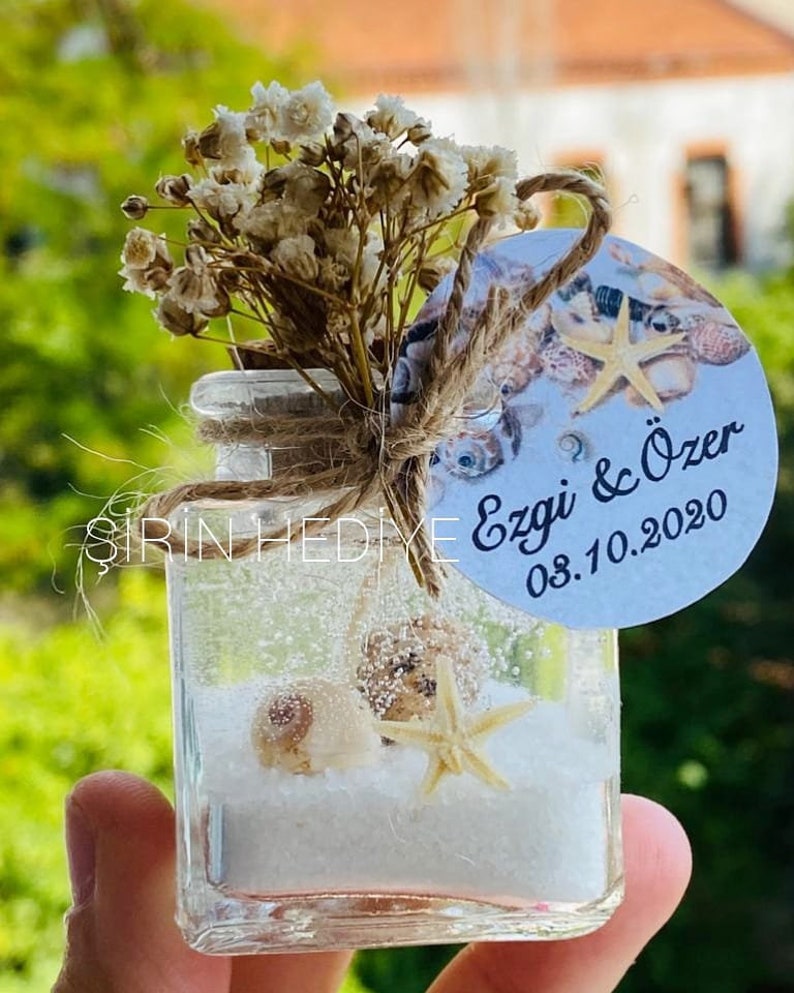 Puede incluir: Un peque&ntilde;o frasco de vidrio lleno de arena blanca y conchas marinas, incluyendo una estrella de mar. El frasco est&aacute; decorado con un lazo de cuerda marr&oacute;n y una etiqueta redonda blanca con el texto "Ezgi & &Ouml;zer 08.10.2020".