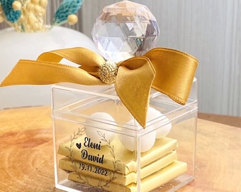 May include: A clear plastic box with a gold ribbon and a crystal top. The box contains white chocolate squares and white candy. The box has a label that says "Eleni David 19.11.2022".