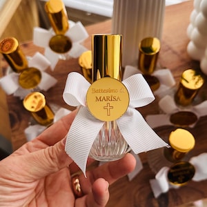 May include: A small, clear glass bottle with vertical ridges, topped with a shiny gold cap. A white grosgrain ribbon is tied around the neck, featuring a circular gold charm engraved with the text Battesimo di MARISA and a cross symbol.