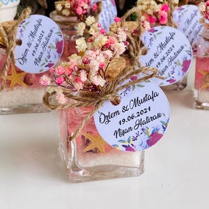 Puede incluir: Peque&ntilde;os frascos de vidrio llenos de arena rosa y una estrella de mar, decorados con flores secas y una etiqueta blanca con el texto "&Ouml;zlem & Mustafa 19.06.2021 Nisan Hatirası".