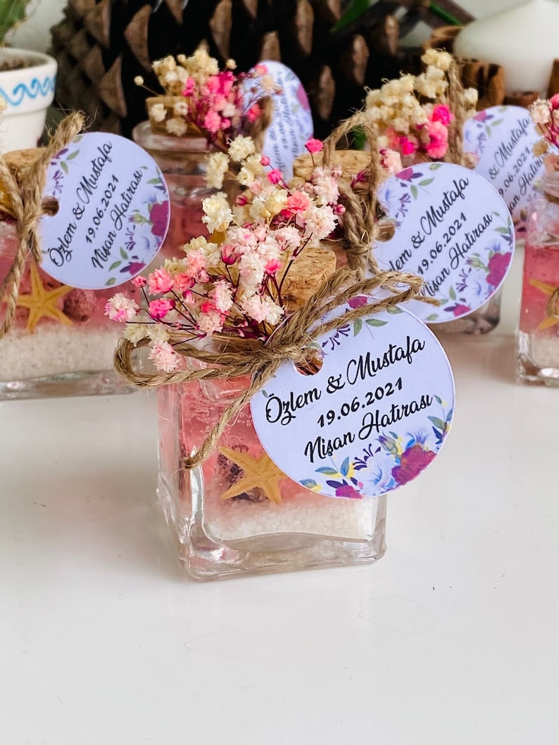 Puede incluir: Peque&ntilde;os frascos de vidrio llenos de arena rosa y una estrella de mar, decorados con flores secas y una etiqueta blanca con el texto "&Ouml;zlem & Mustafa 19.06.2021 Nisan Hatirası".
